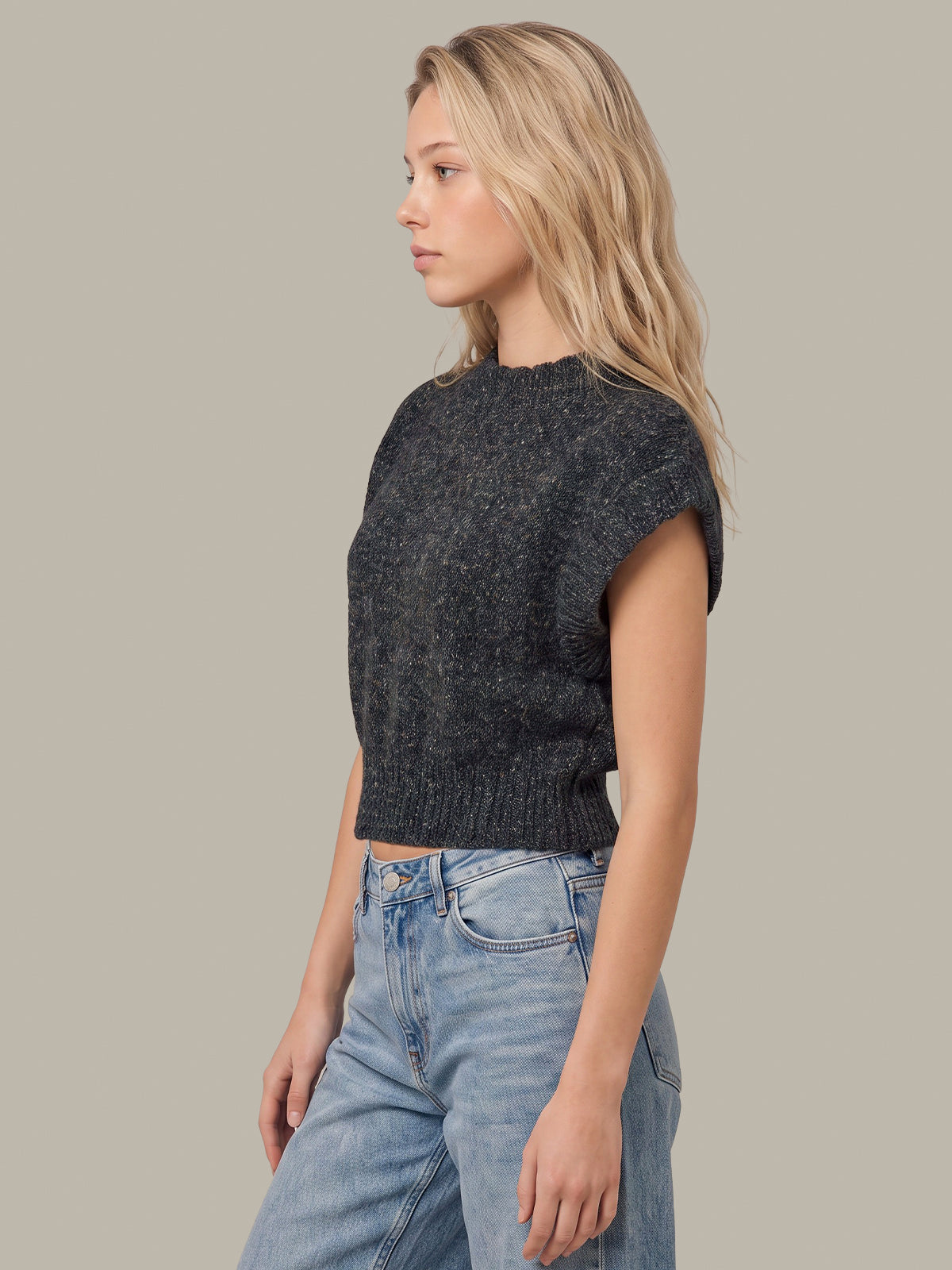 A woman in profile wearing a dark charcoal gray cropped knit sweater vest and light blue jeans, set against a solid warm gray studio background.