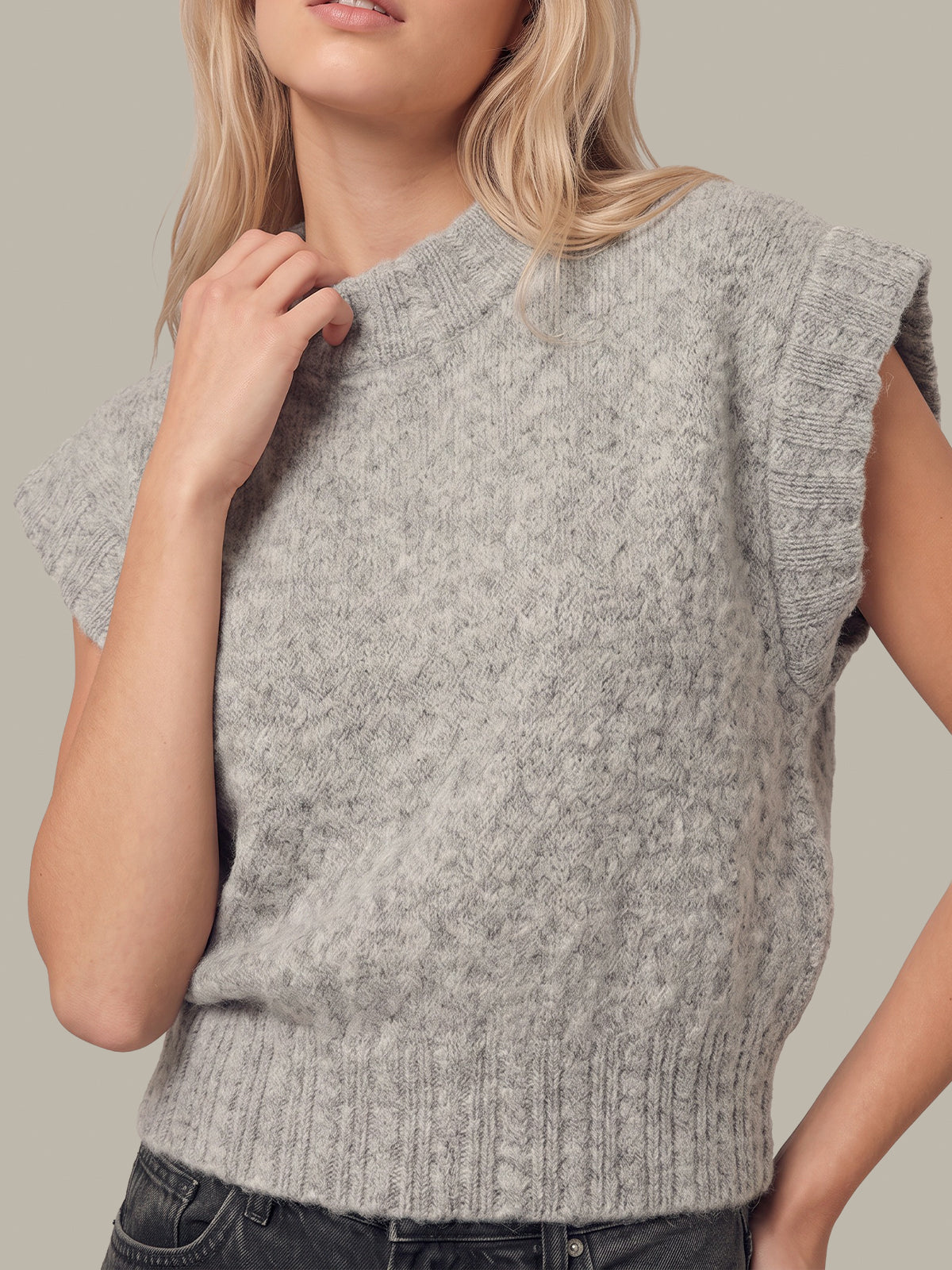 A close-up of a woman wearing a light gray ribbed knit sweater vest with textured cable-like patterns and ruffled sleeve edges, paired with dark jeans against a solid neutral background.