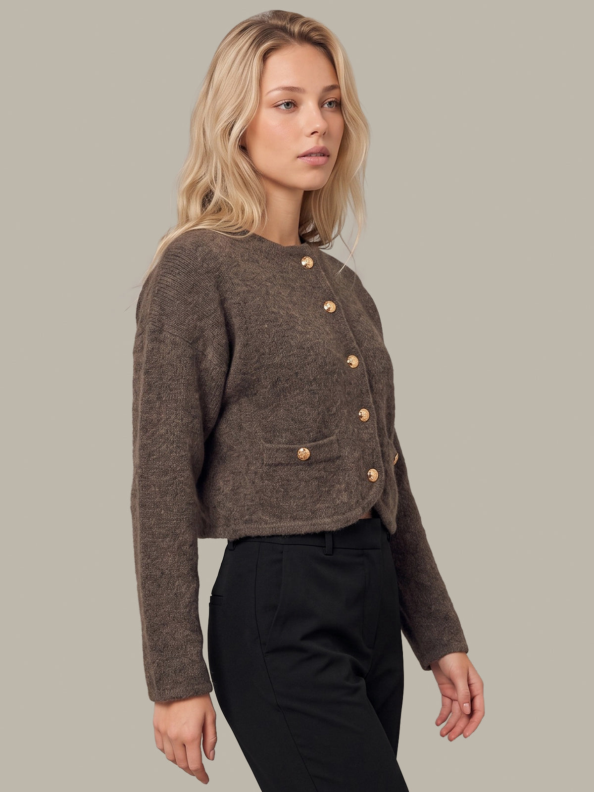 A woman modeling a cropped, textured brown cardigan with gold buttons, paired with black trousers, against a solid light gray background.
