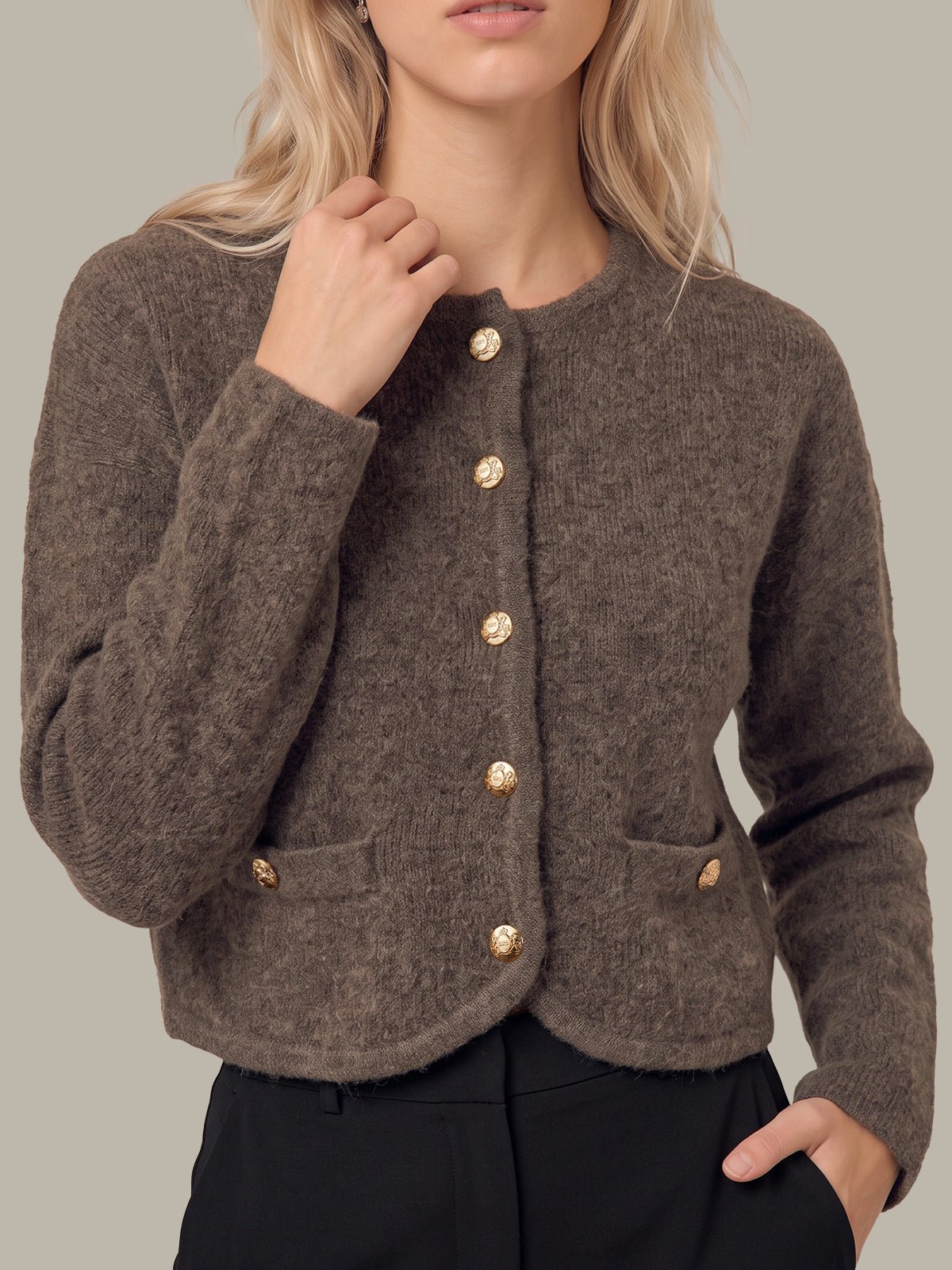 A woman wearing a cropped, fuzzy brown knit cardigan with gold buttons and front pockets, paired with black trousers, against a solid neutral background.