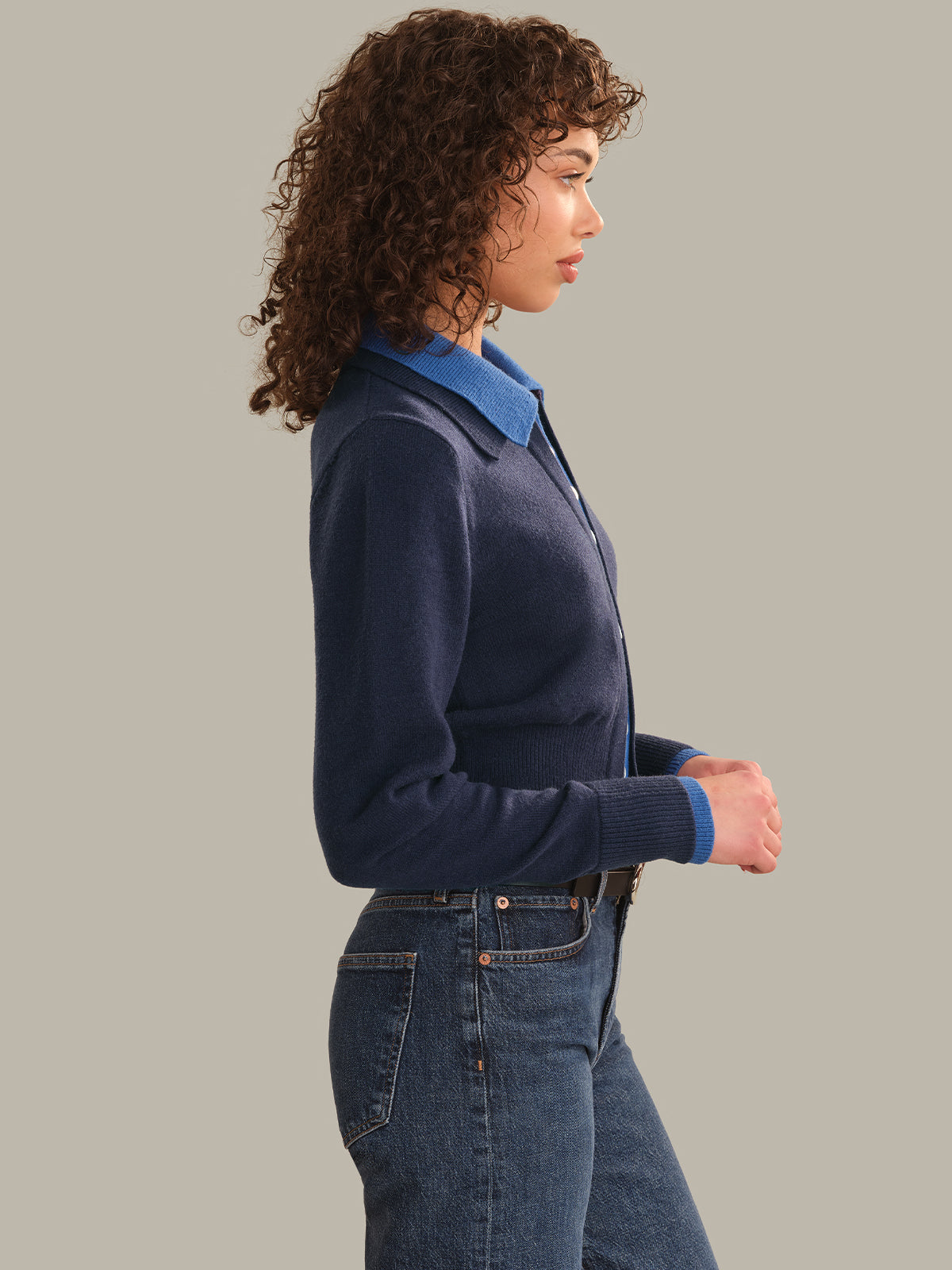 A woman shown in side profile wearing a navy blue knit cardigan over a blue collared shirt, paired with blue jeans, against a solid light gray studio background.