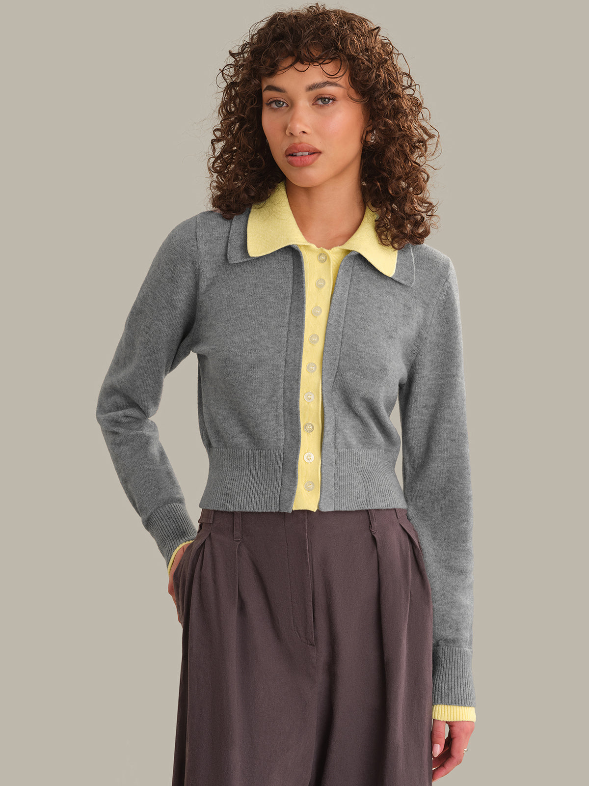 A woman with curly brown hair wearing a gray knit cardigan over a light yellow collared button-up shirt, paired with a dark brown skirt, against a solid light gray background.