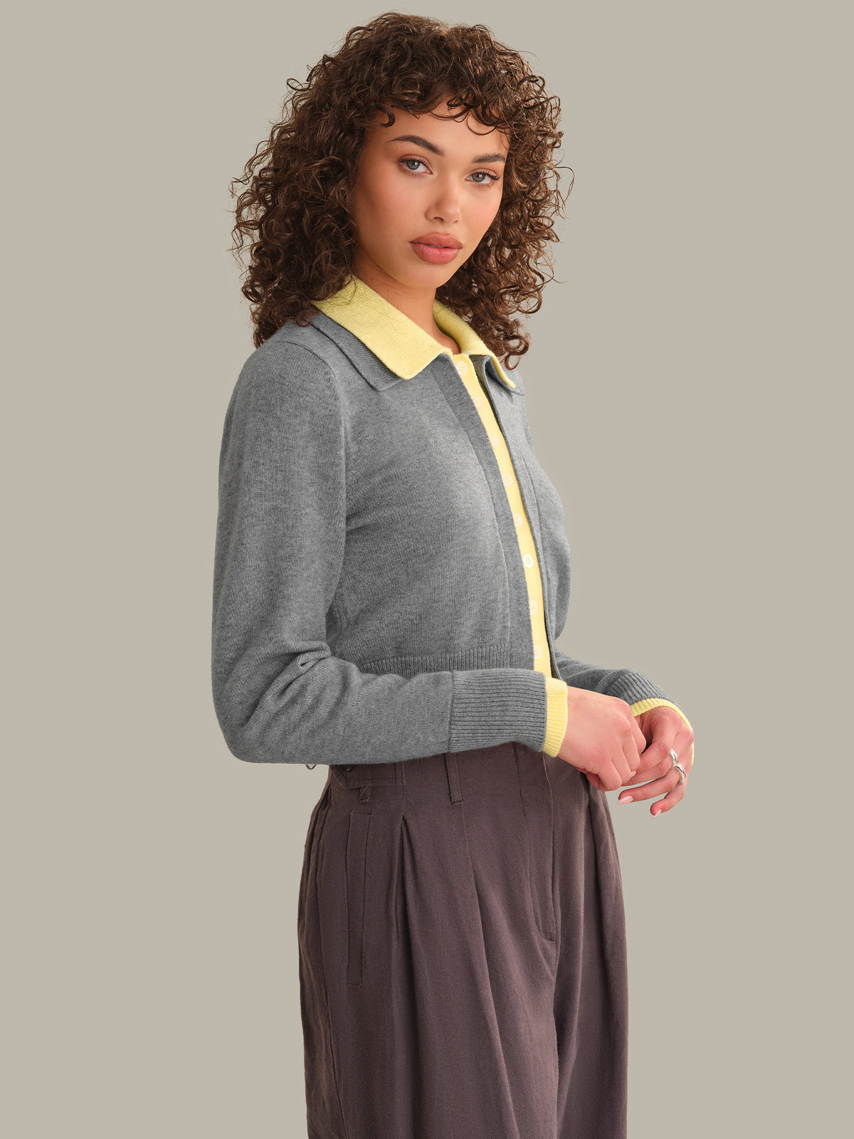 A woman modeling a gray knit cardigan with a contrasting light yellow collar and front placket, paired with dark brown pleated trousers, against a solid light gray background.