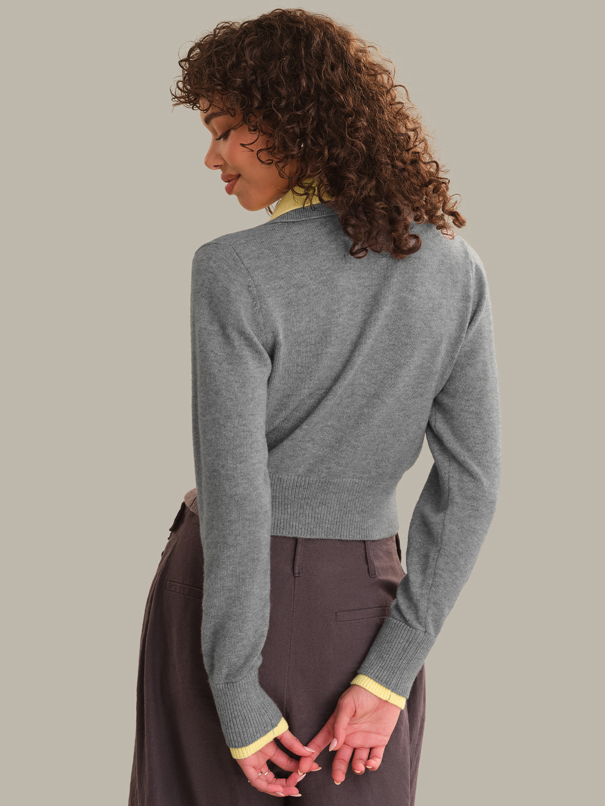 A woman with curly brown hair is shown from the back wearing a heather gray ribbed knit cardigan with yellow contrast trim at the collar and cuffs, paired with dark brown trousers, against a solid light gray background.