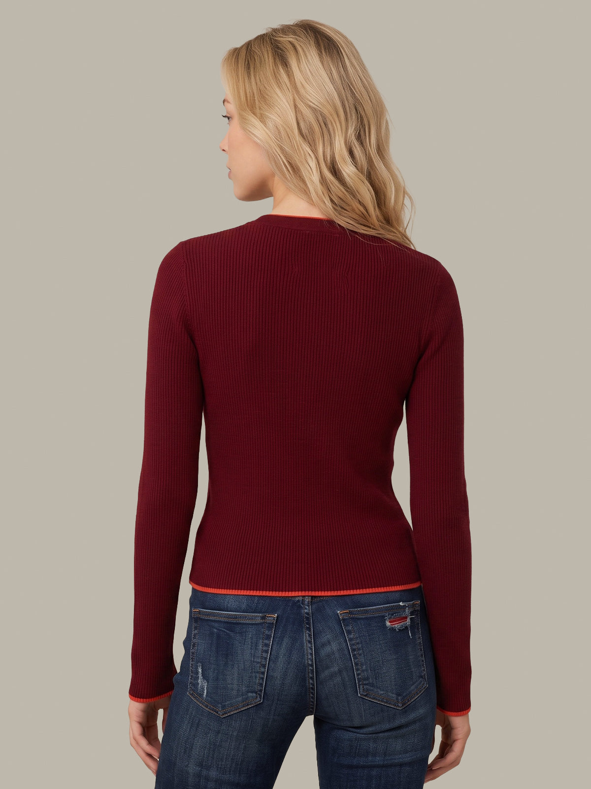 A woman viewed from the back wearing a fitted ribbed long-sleeve sweater in deep burgundy and blue jeans, against a plain warm gray studio background.