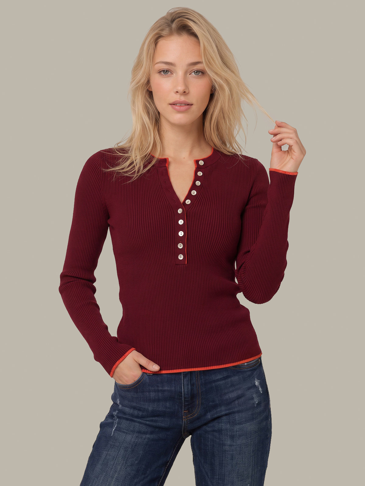 A woman modeling a ribbed long-sleeve henley top in deep burgundy with white buttons and orange-red trim, paired with blue jeans against a solid light gray background.