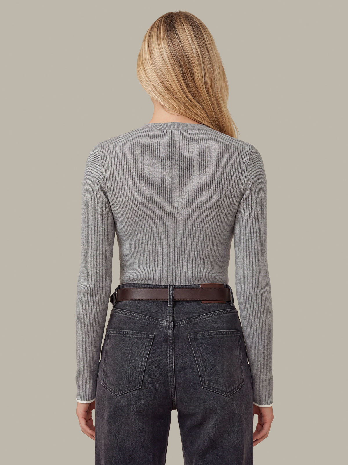 A woman shown from the back wearing a fitted light gray ribbed long-sleeve top, dark gray jeans, and a brown belt against a plain warm gray background.