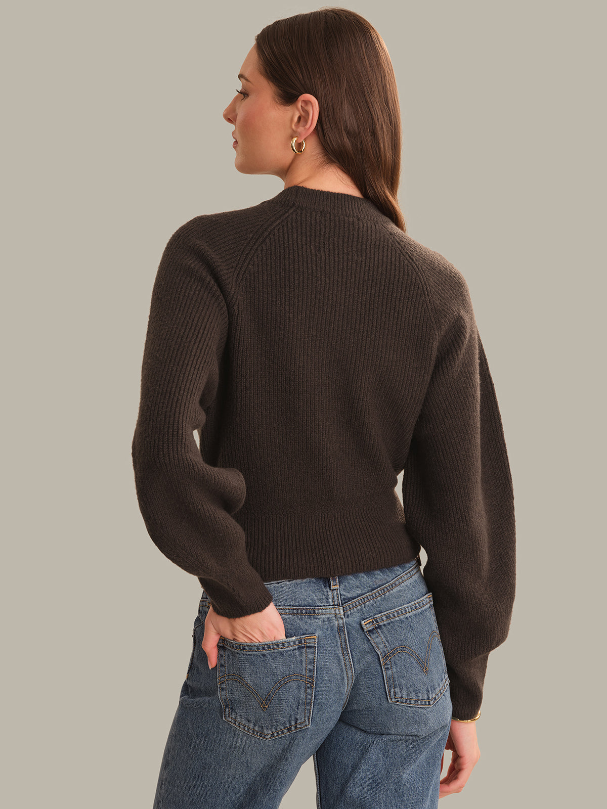 A woman shown from the back wearing a dark brown ribbed knit sweater and blue jeans against a solid light gray background.