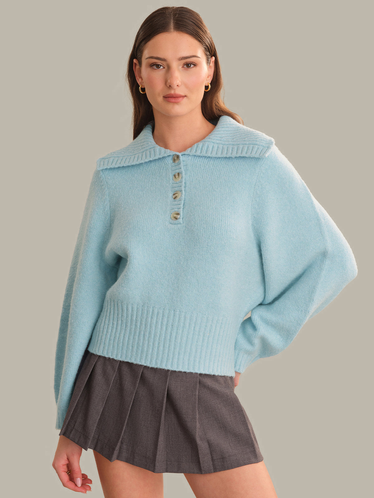 A woman modeling a light blue knit sweater with a wide collar and three decorative buttons, paired with a dark gray pleated mini skirt, against a solid taupe background.
