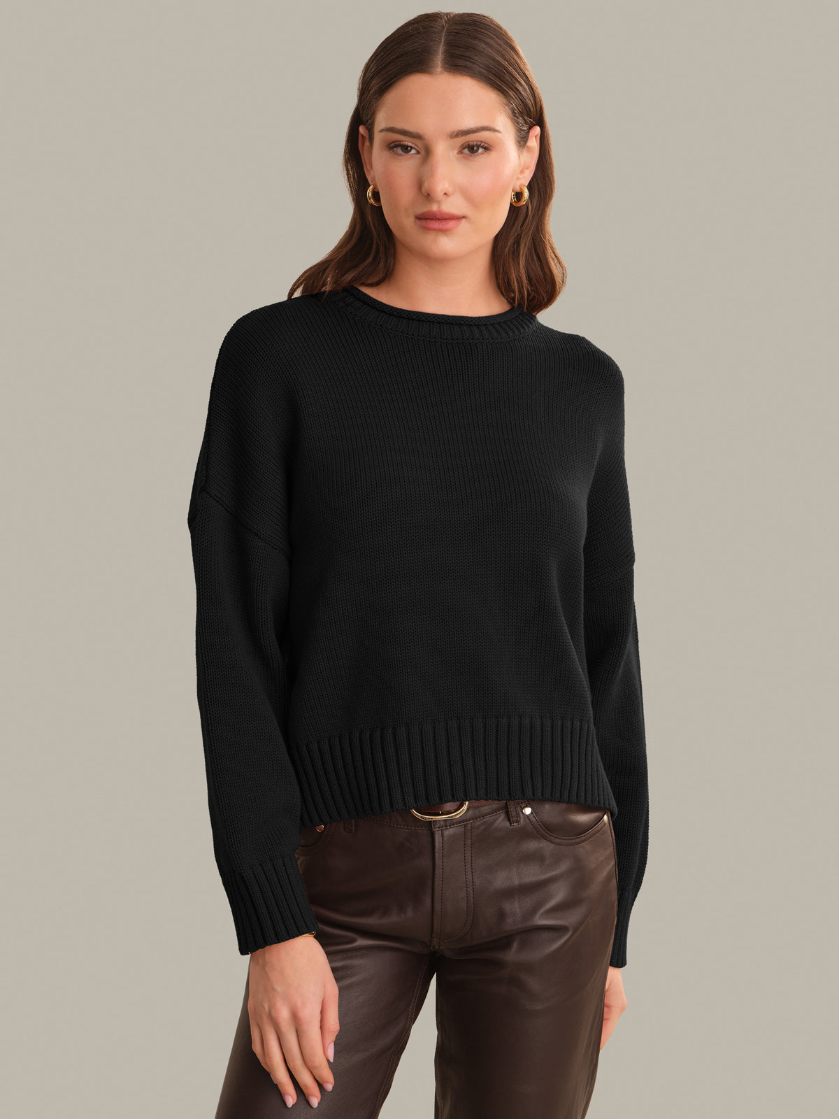 A woman modeling a black long-sleeve knit sweater with a crew neck and ribbed hem, paired with dark brown pants, against a solid light gray background.