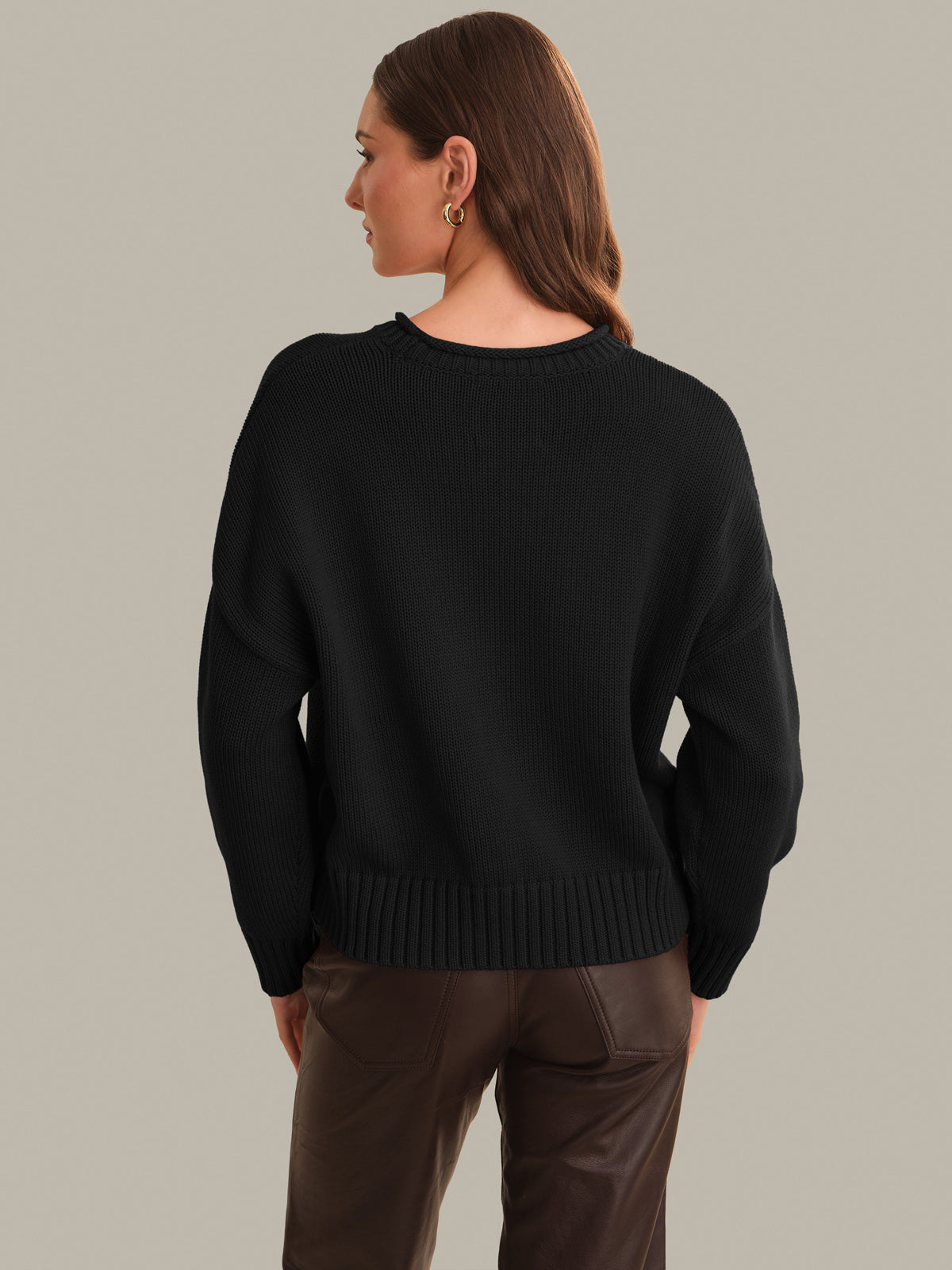 A woman shown from the back wearing a black knit sweater and brown pants against a plain light gray studio background.