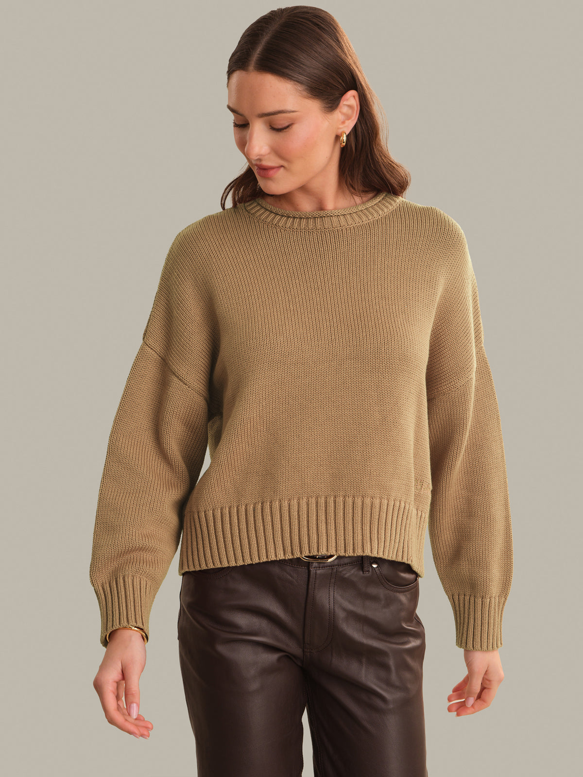 A woman wearing a cropped, ribbed knit sweater in camel beige with dark brown faux-leather pants, standing against a solid light gray background.