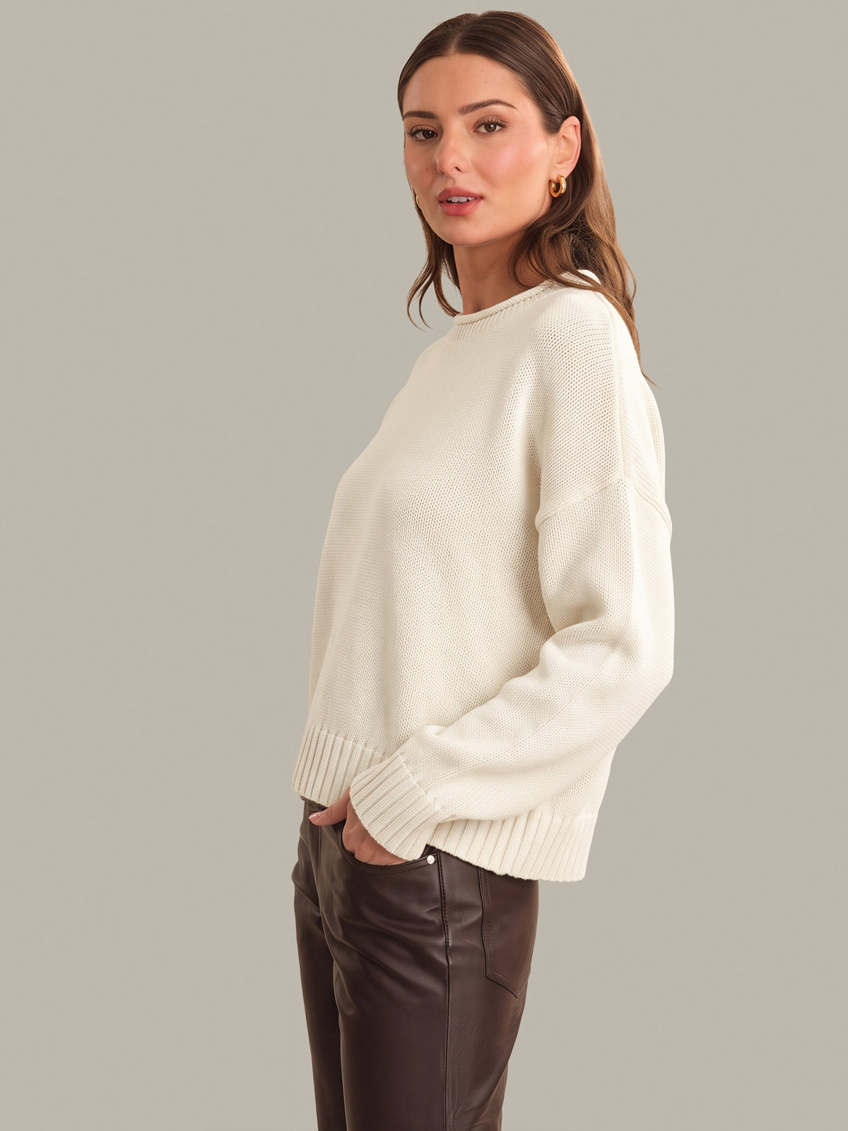 A woman modeling an oversized cream knit sweater with ribbed cuffs and hem, paired with dark brown pants, against a solid light gray studio background.
