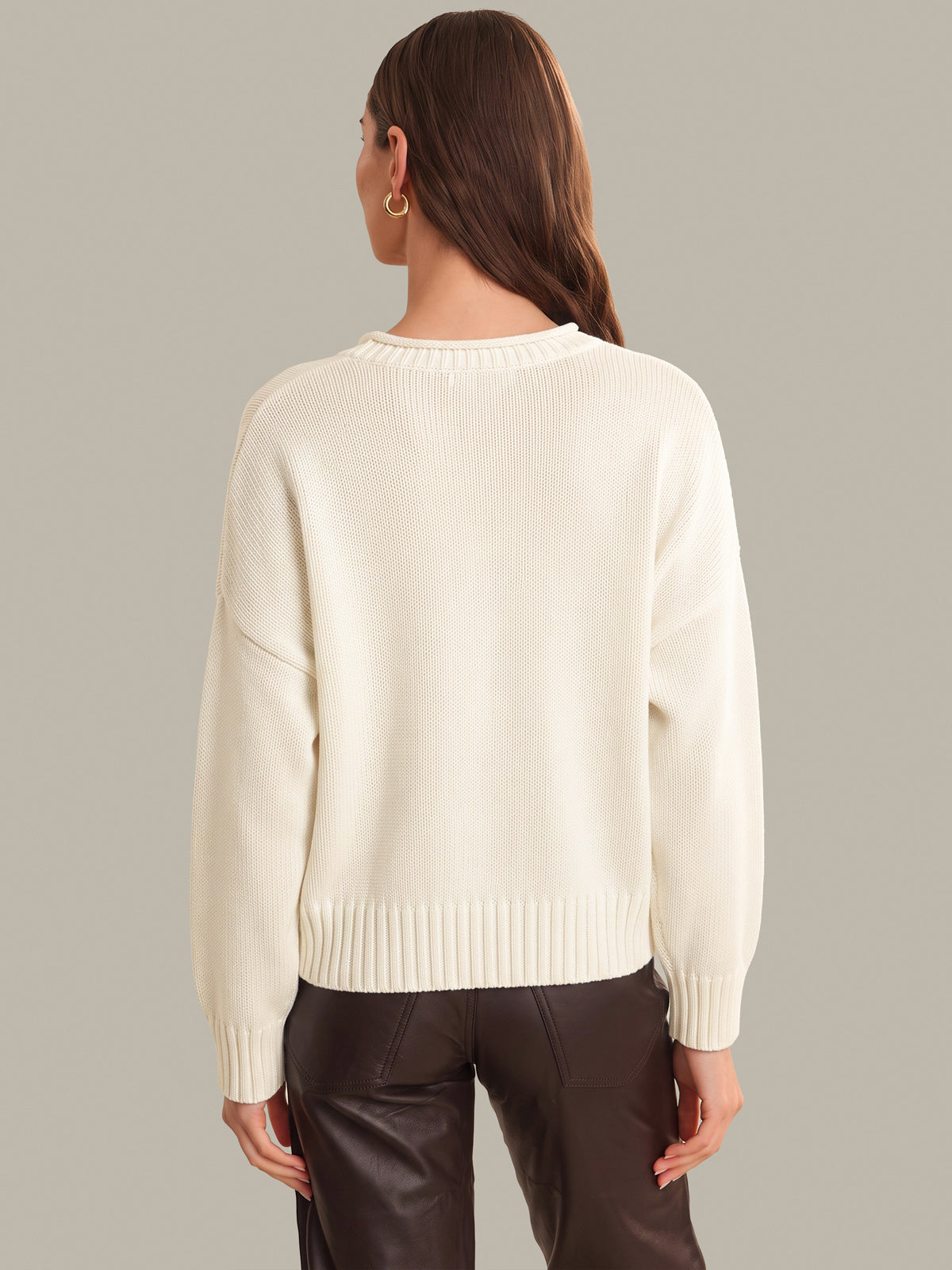 A woman shown from the back wearing a cream-colored ribbed knit sweater and dark brown pants, set against a solid light gray studio background.