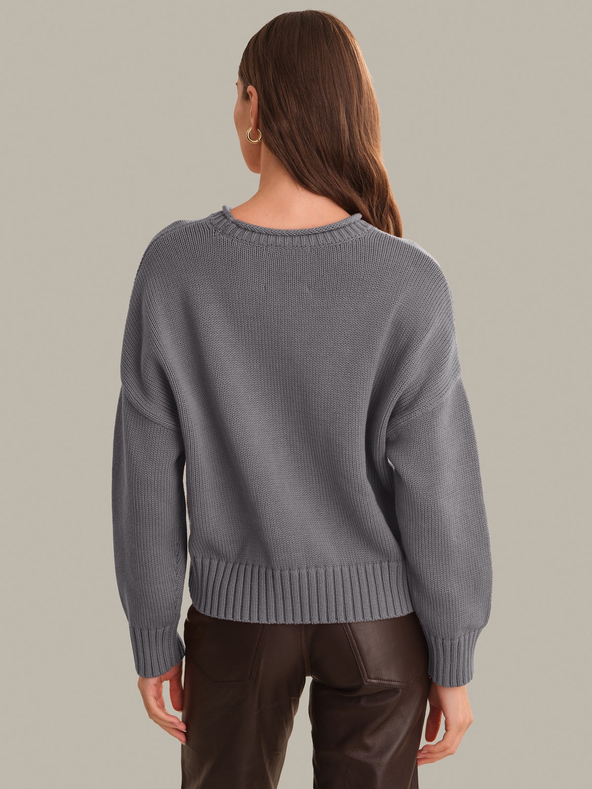 A woman viewed from the back wearing a gray ribbed knit sweater and dark brown pants, standing against a plain warm gray studio background.