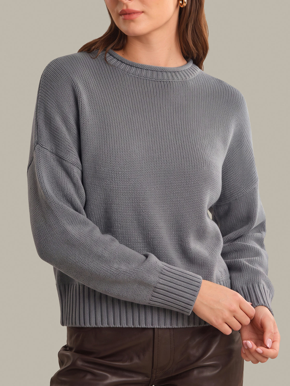 A woman wearing a gray ribbed knit sweater with long sleeves and a crew neck, paired with dark brown leather pants, against a solid light gray background.