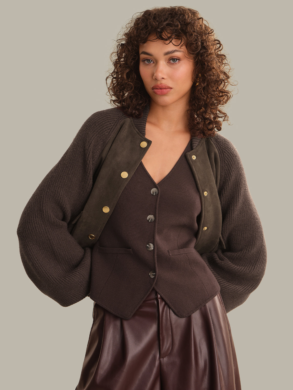 A model with curly brown hair wearing a dark brown ribbed knit cardigan with gold-tone snap buttons over a matching brown vest and a dark brown faux-leather skirt, set against a solid light gray background.