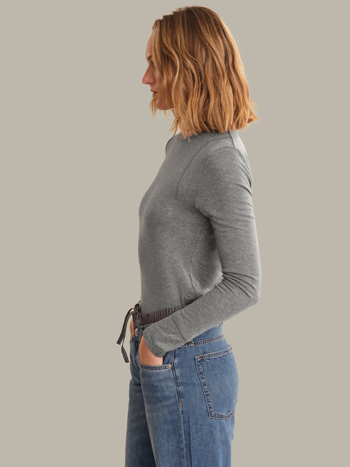 A woman shown in side profile wearing a fitted heather gray long-sleeve top and blue jeans against a plain warm gray studio background.