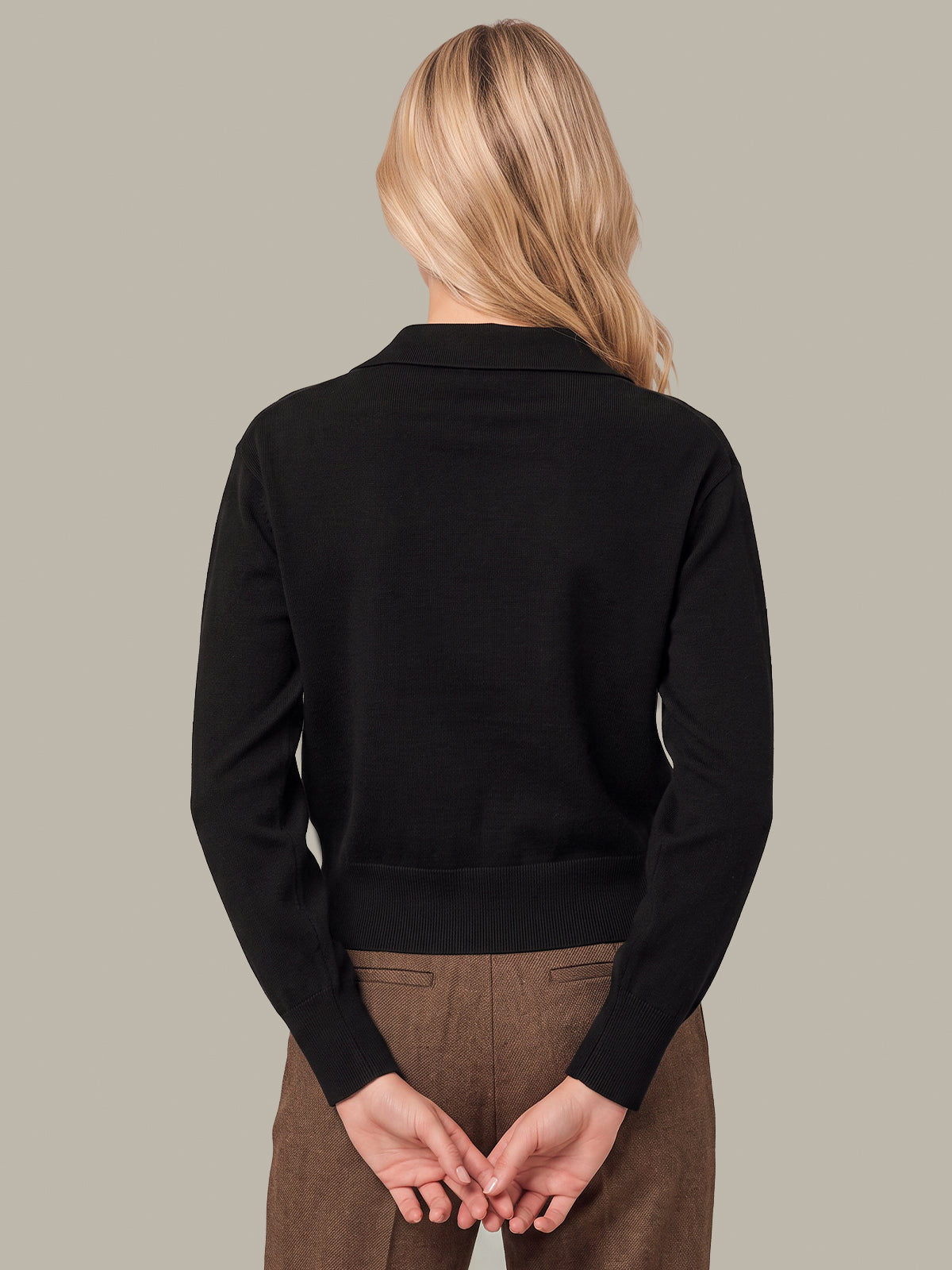 A woman viewed from the back wearing a black long-sleeve knit top with a collar and brown trousers, standing against a solid light gray background.