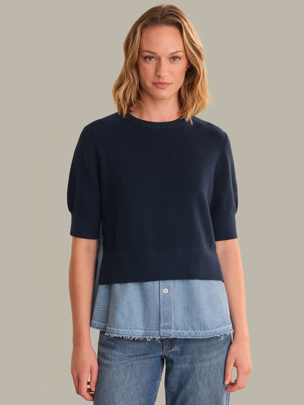 Woman wearing a navy sweater over a denim shirt with a plain background