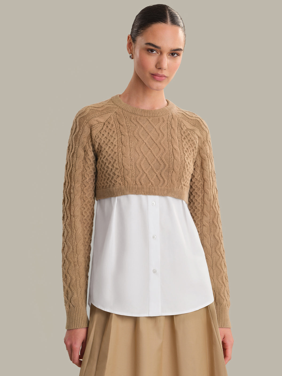 Woman wearing a beige cable knit sweater over a white shirt with a beige skirt on a plain background