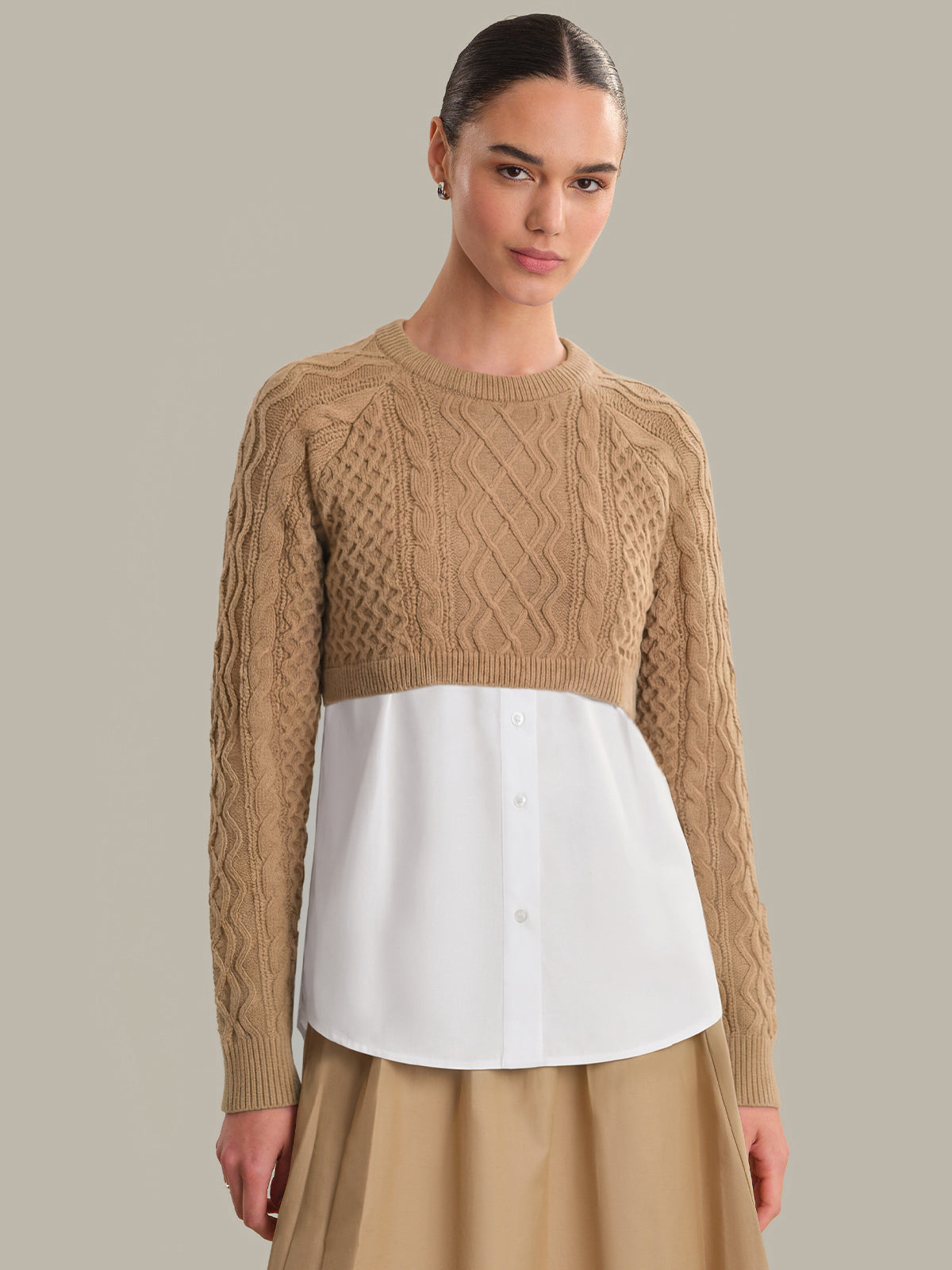 Woman wearing a beige cable knit sweater over a white shirt with a beige skirt on a gray background
