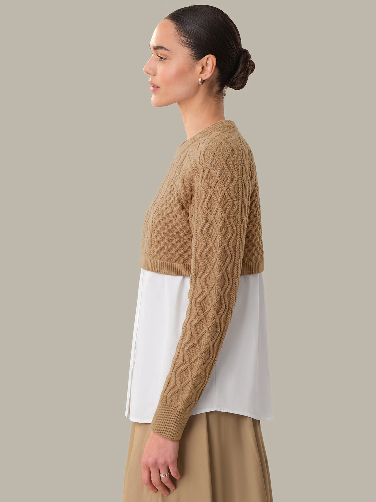 Woman wearing a brown cable knit sweater over a white shirt with a beige skirt on a neutral background