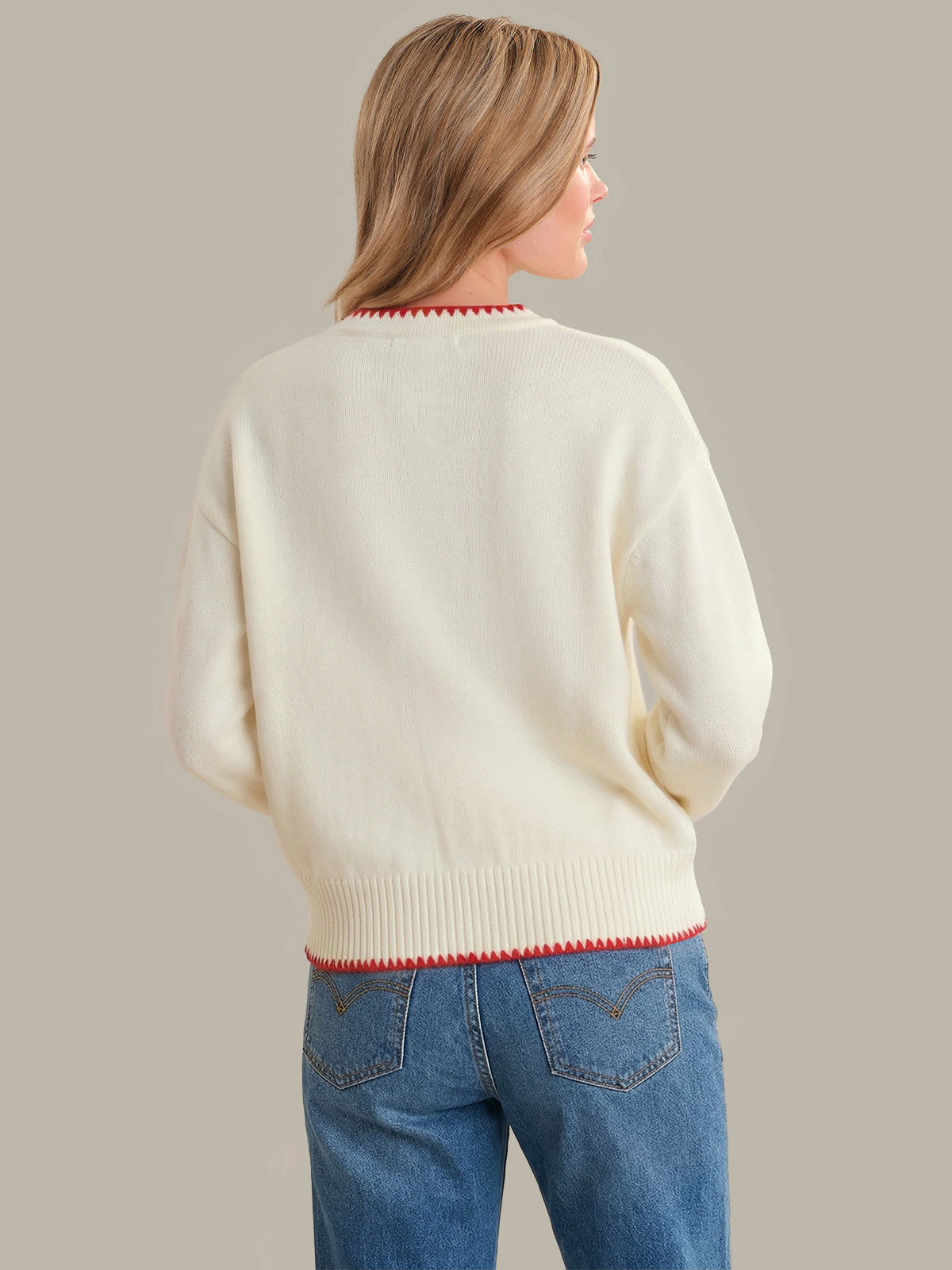 Woman wearing a cream sweater with red trim and blue jeans on a plain background