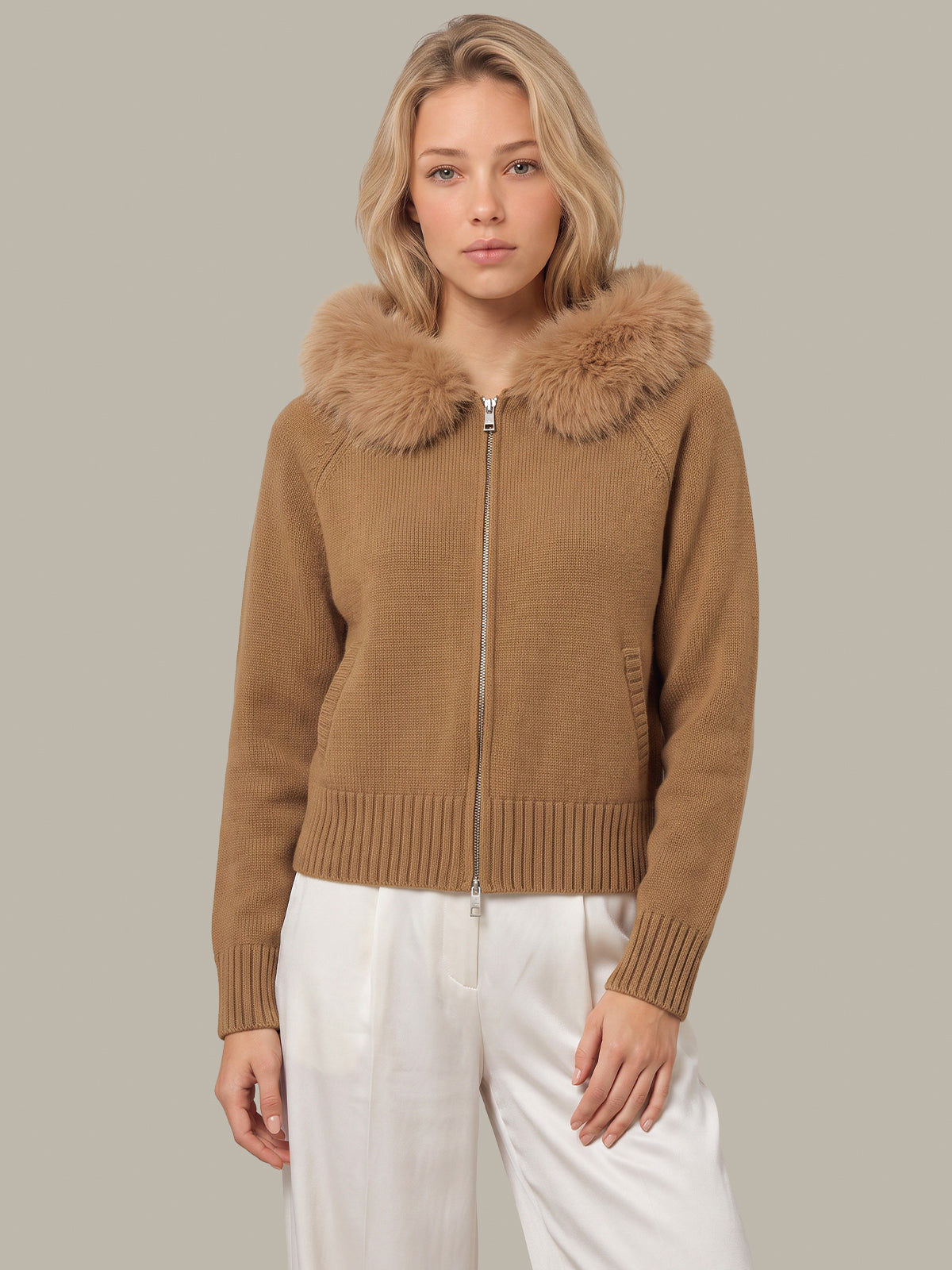 A woman wearing a cropped camel brown knit zip-up jacket with a large fluffy faux-fur collar, paired with white trousers, against a solid light gray background.