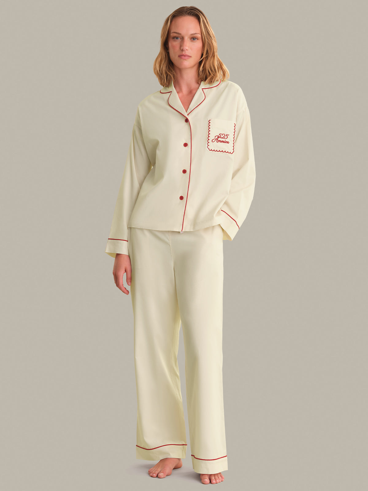 Woman wearing a cream pajama set with red piping on a gray background