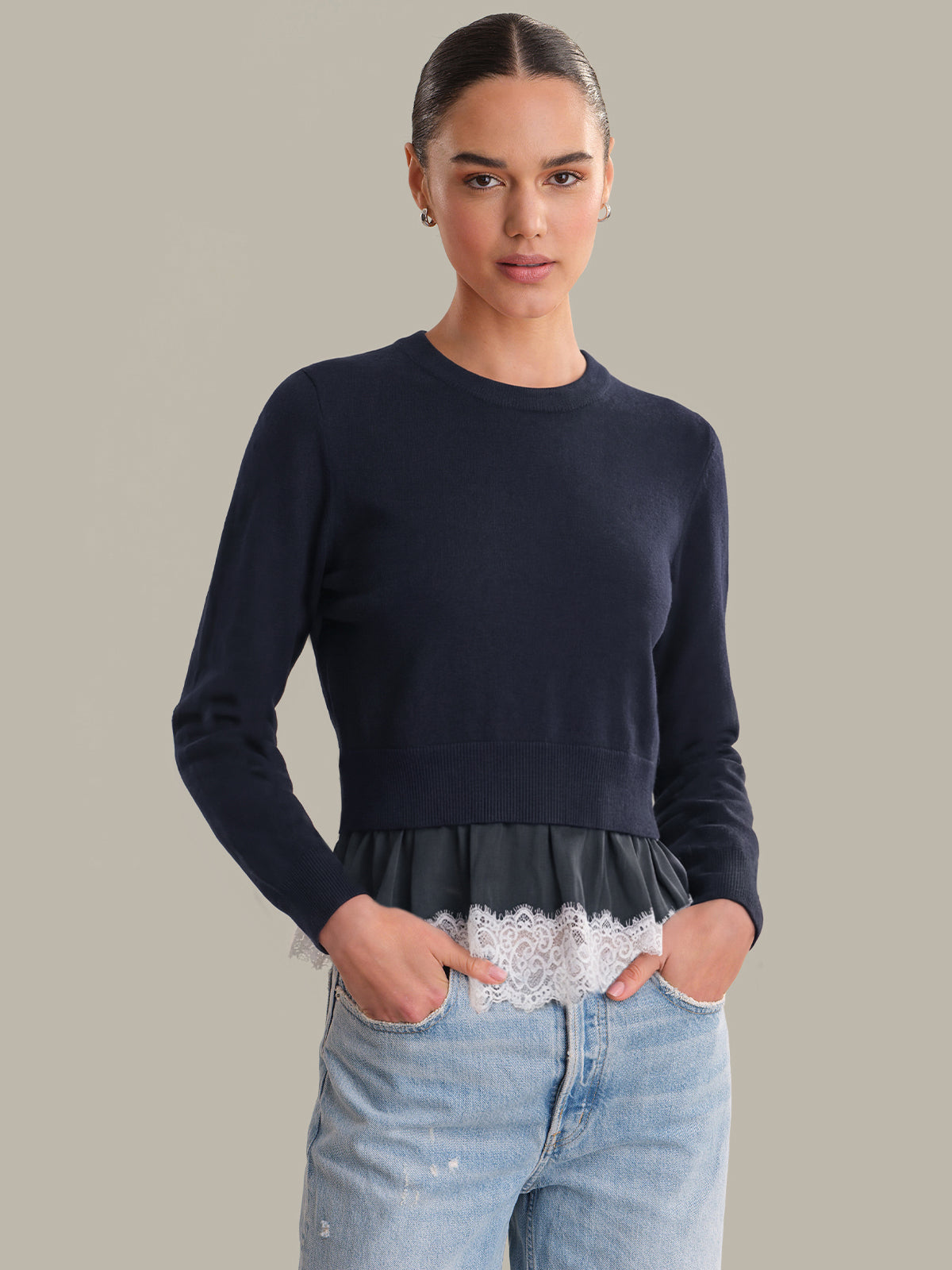 Woman wearing a navy sweater with lace trim and light blue jeans on a neutral background
