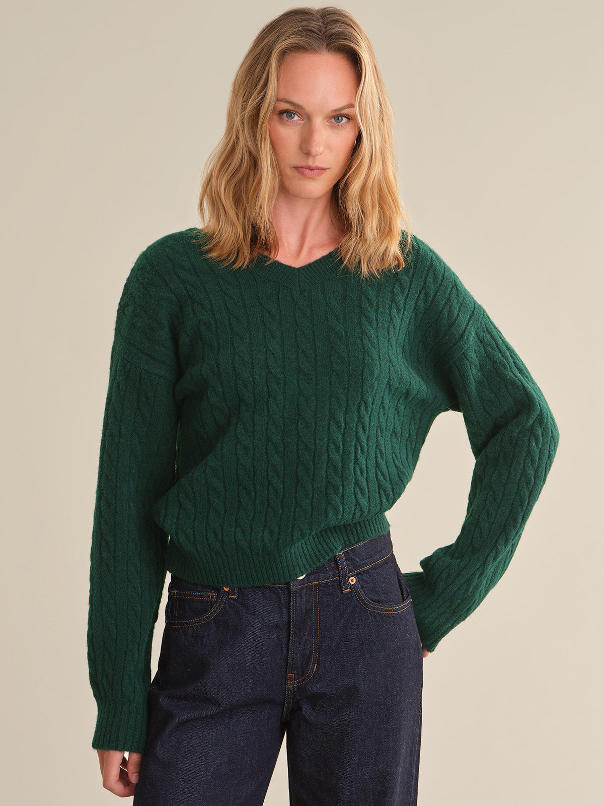 A woman wearing a dark green cable-knit crewneck sweater and dark blue jeans, standing against a solid light beige background.