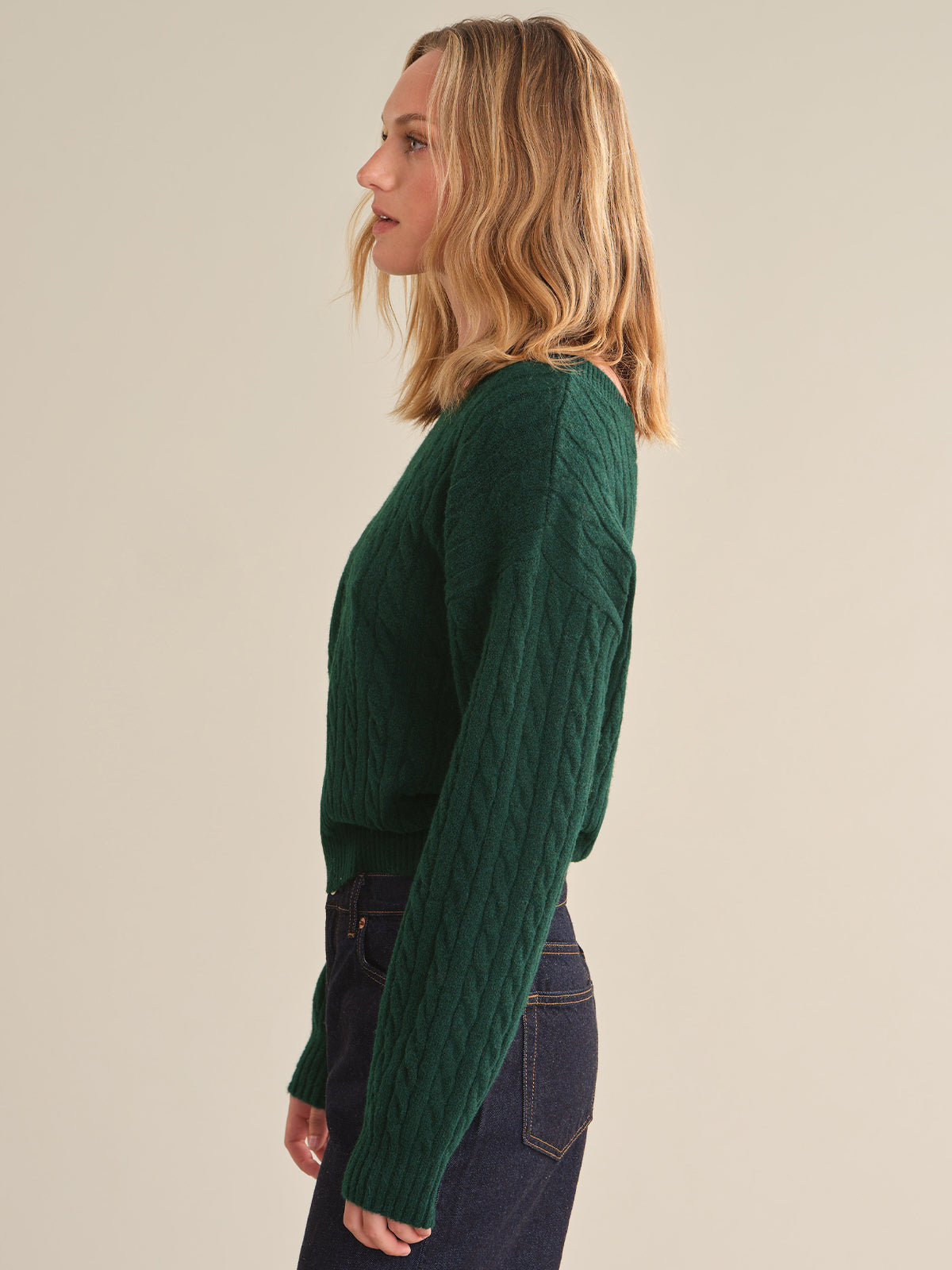 A woman in side profile wearing a dark green cable-knit sweater and dark blue jeans, standing against a plain beige studio background.
