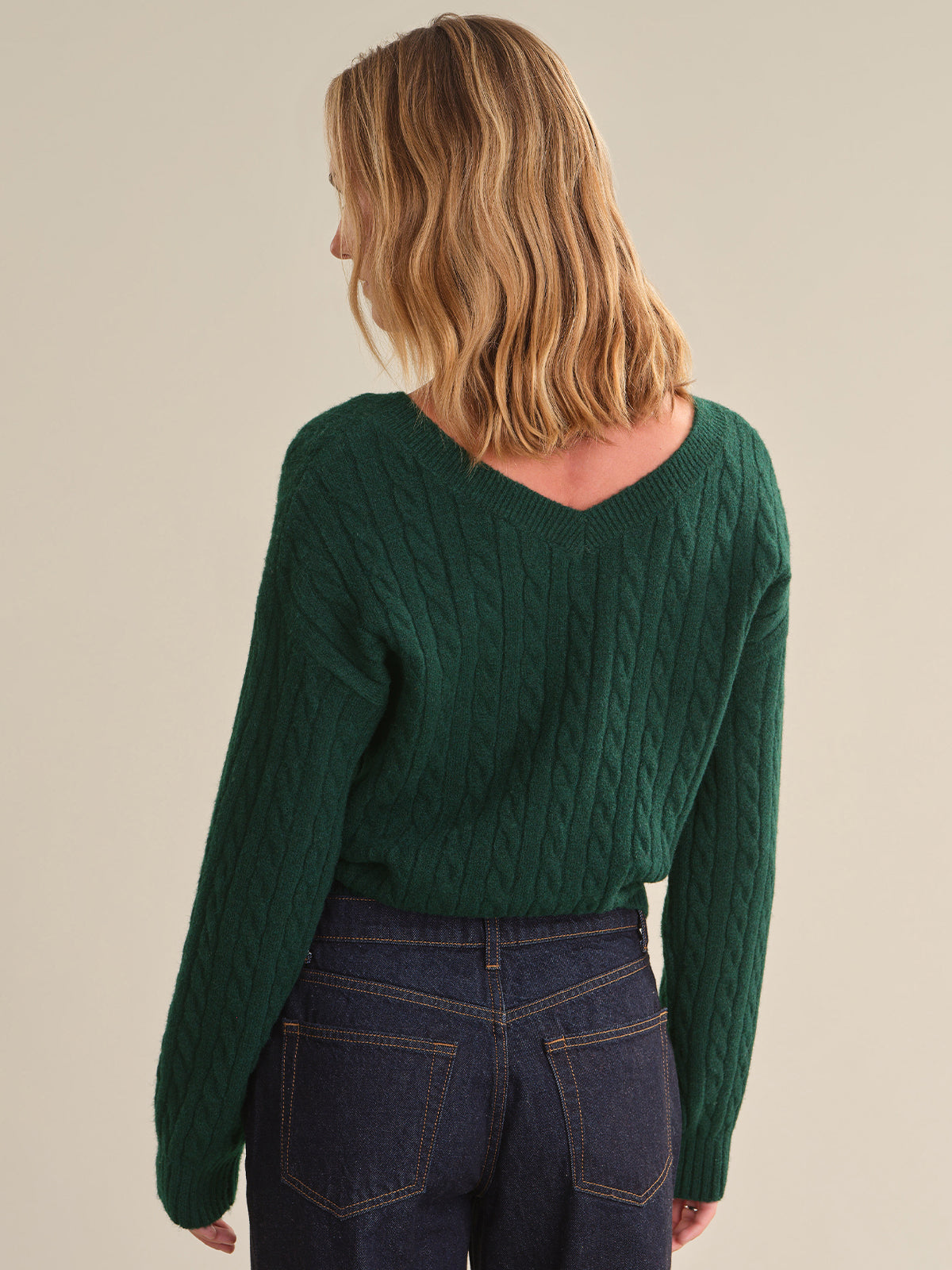 A woman shown from the back wearing a dark green cable-knit sweater and dark blue jeans, against a plain beige studio background.