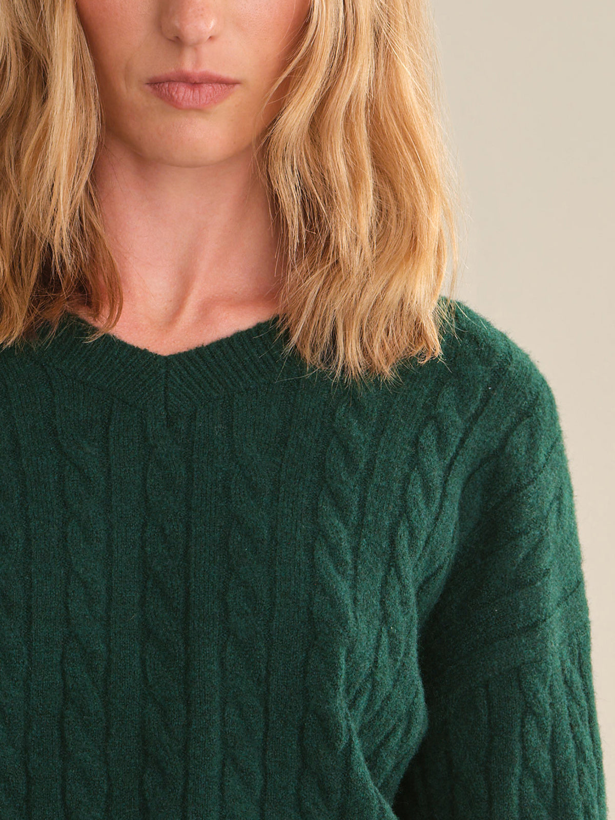 Close-up of a woman wearing a dark green cable-knit sweater, showing the texture of the knit and her blonde hair against a neutral beige background.
