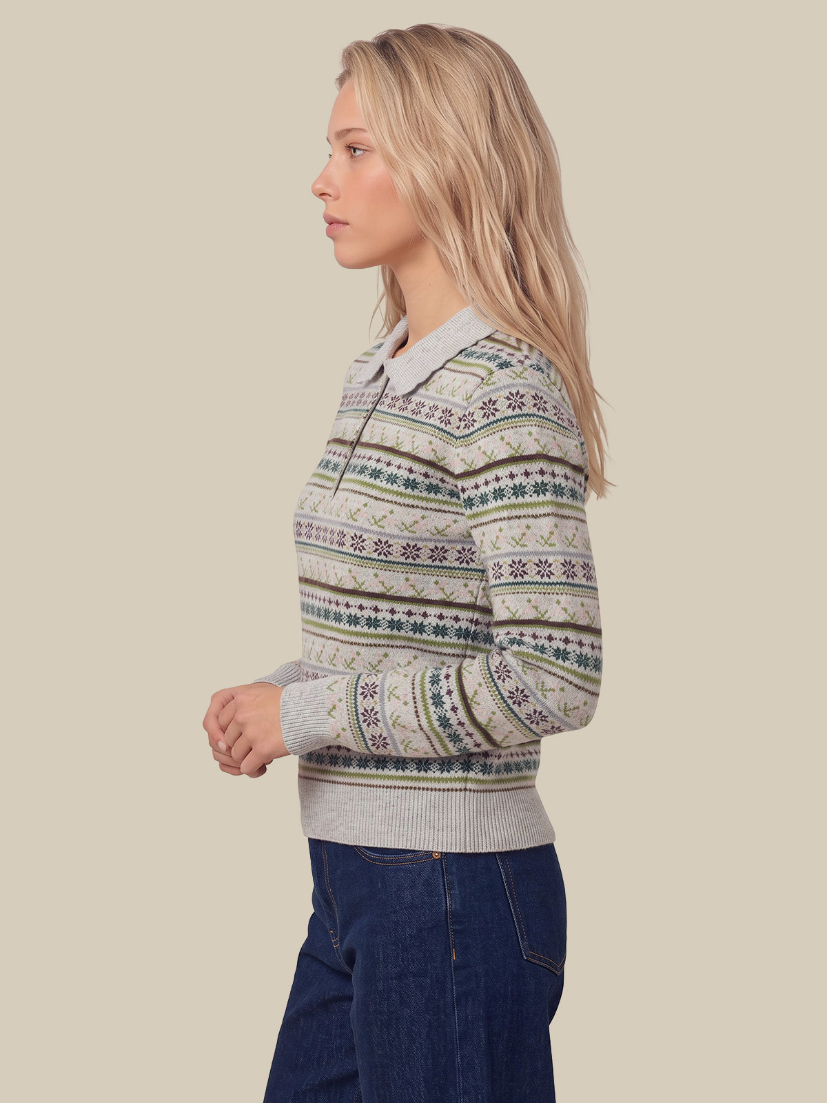 A woman in profile wearing a cream knit sweater with green and blue Nordic Fair Isle patterns, paired with dark blue jeans, against a solid beige background.