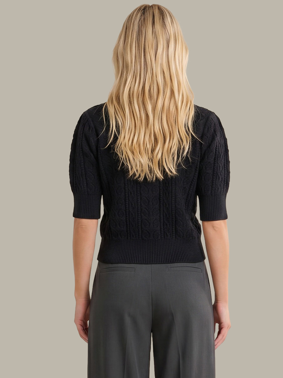 A woman viewed from the back wearing a dark navy cable-knit short-sleeve cropped sweater and dark gray trousers, set against a solid light gray background.