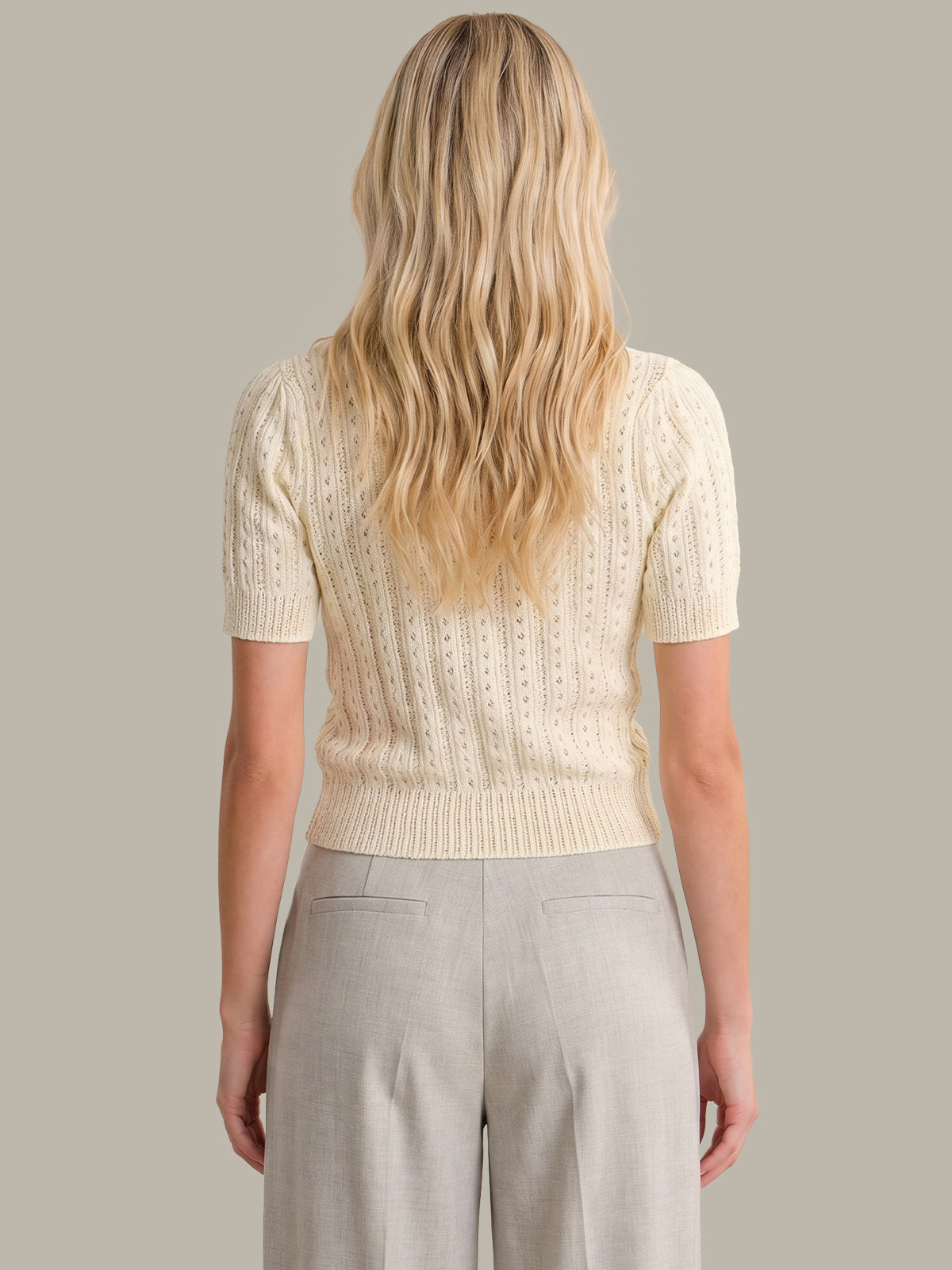 A woman shown from the back wearing a cream short-sleeve cable-knit top and light beige trousers, against a solid taupe background.