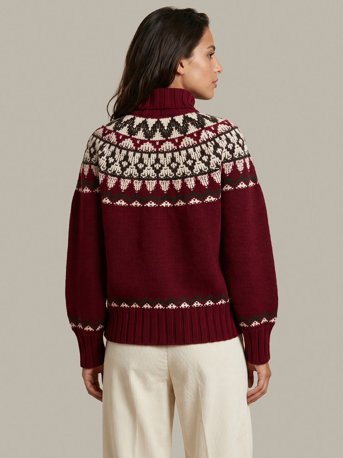 A woman shown from the back wearing a burgundy turtleneck sweater with a cream and black Fair Isle pattern across the shoulders and upper back, paired with cream trousers, against a solid light gray background.