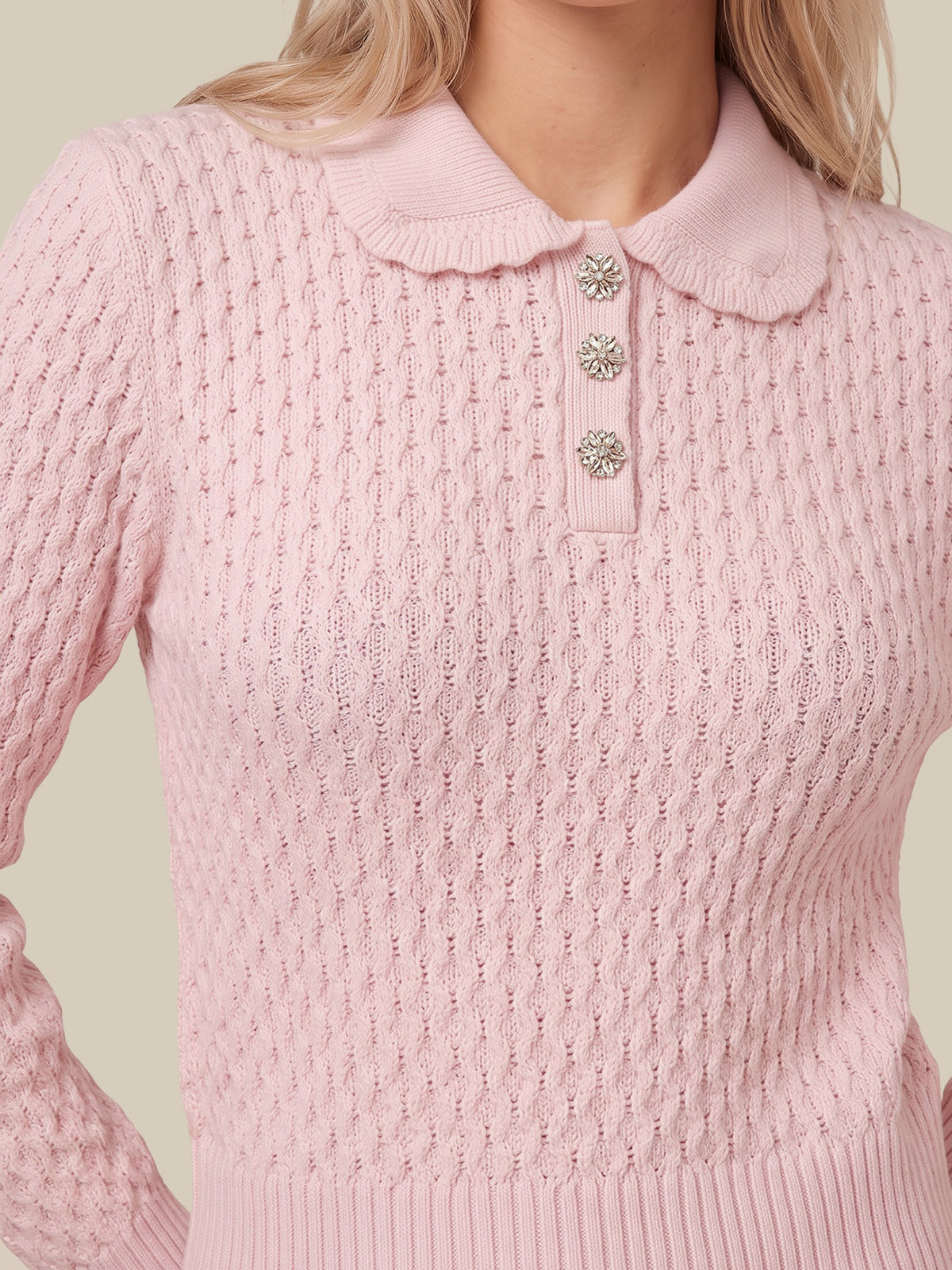 A close-up of a woman wearing a light pink cable-knit sweater with a Peter Pan collar and three decorative silver-tone buttons.