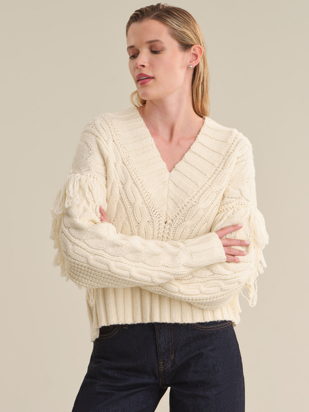 A woman wearing a cream-colored cable-knit sweater with fringe details on the sleeves, paired with dark blue jeans, against a solid beige background.
