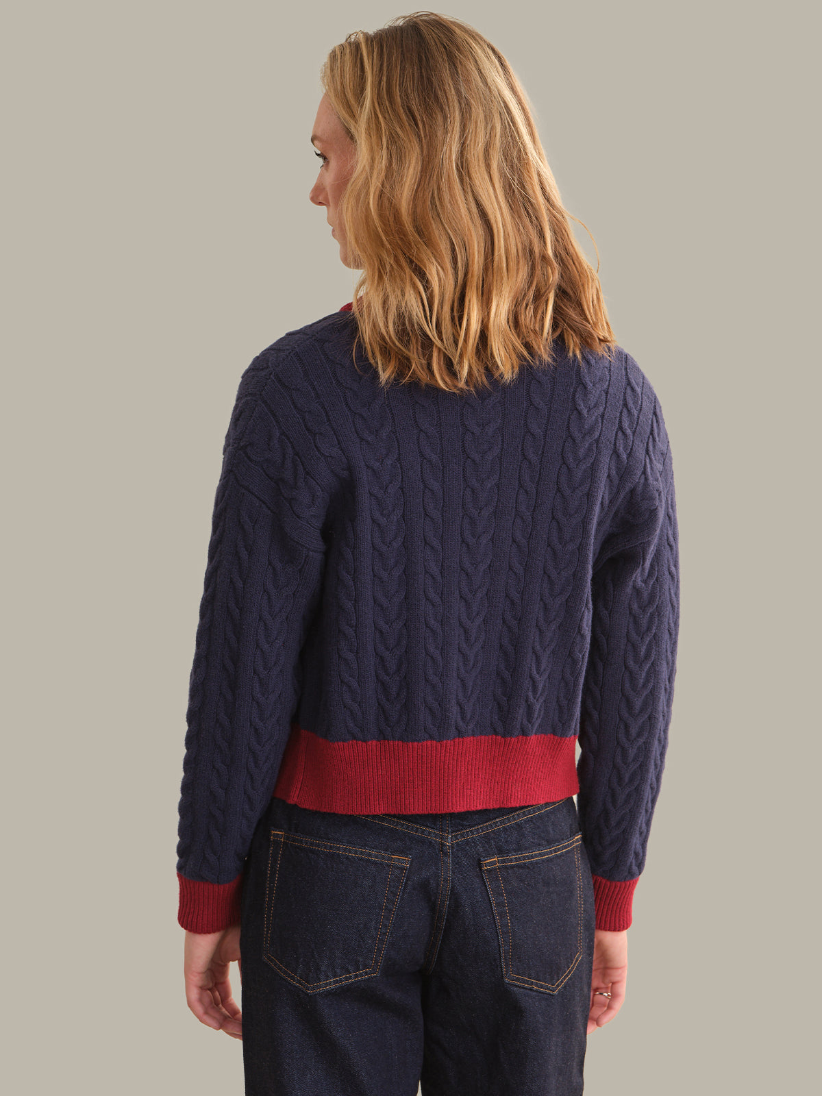 A woman shown from the back wearing a navy blue cable-knit sweater with a contrasting red hem and cuffs, paired with dark blue jeans, against a solid light gray background.