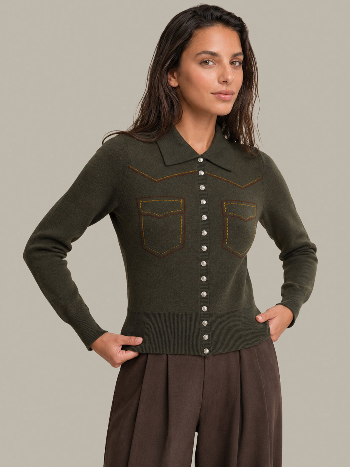A woman modeling a dark olive green knit cardigan with brown western-style stitching and button details, paired with brown pants, against a solid light gray background.