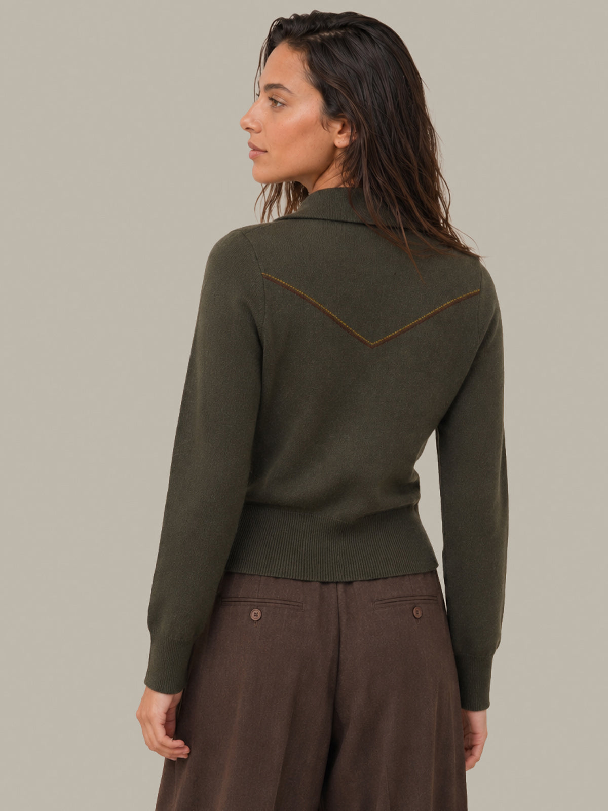A woman shown from the back wearing an olive green knit sweater with a brown chevron detail on the upper back, paired with brown pants, against a solid light gray background.