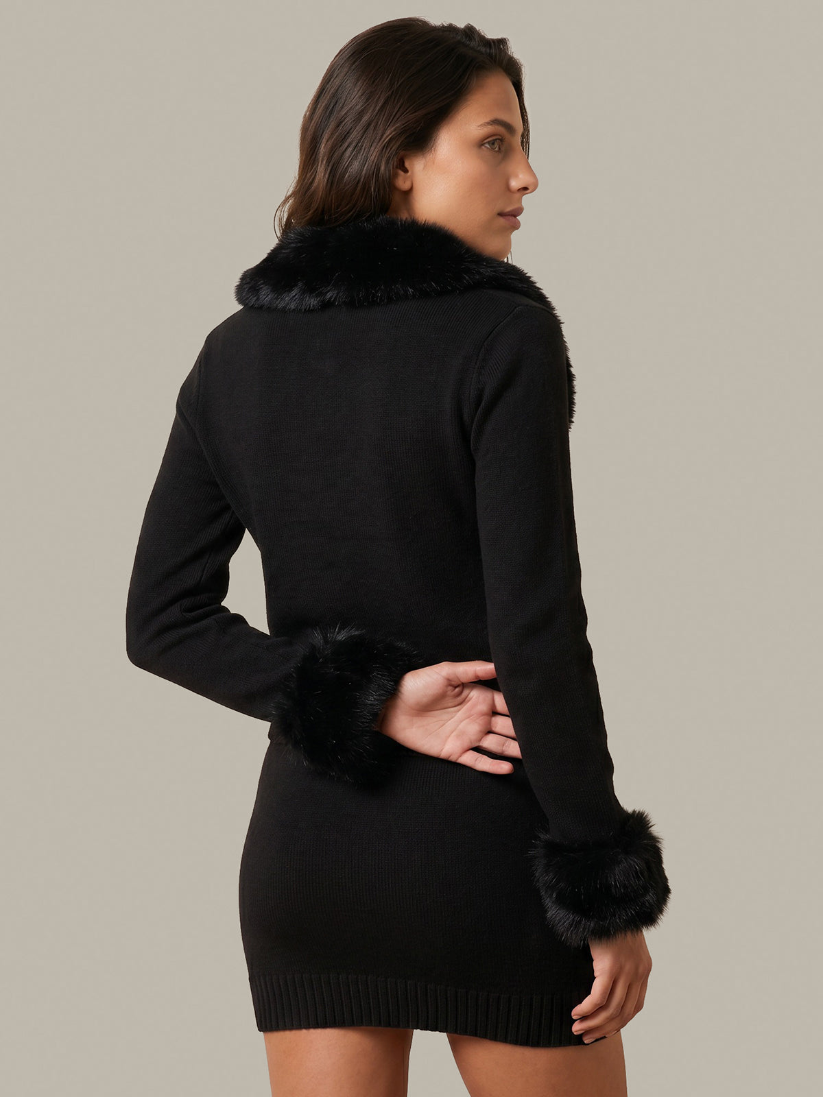 A woman wearing a black knit mini dress with black faux-fur trim around the collar and cuffs, shown from a three-quarters back view against a solid light gray background.