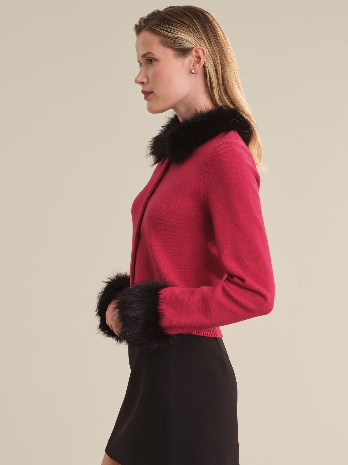 A woman shown in side profile wearing a bright pink knit cardigan with black faux-fur trim at the collar and cuffs, paired with a black skirt, against a plain beige studio background.