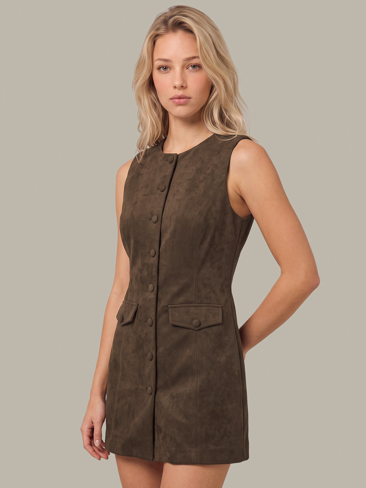 A blonde woman modeling a sleeveless, dark brown faux-suede mini dress with a button-down front and flap pockets, set against a solid light gray background.