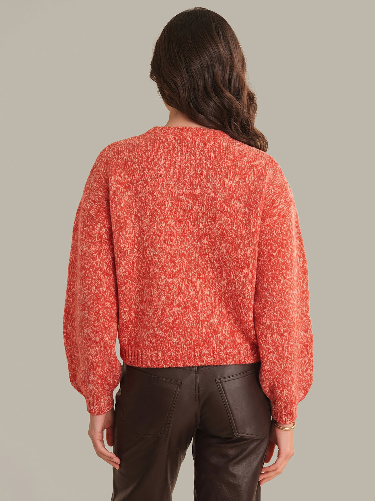 A woman viewed from the back wearing a textured, marled coral-orange knit sweater with balloon sleeves and dark brown faux-leather pants, set against a solid light gray background.