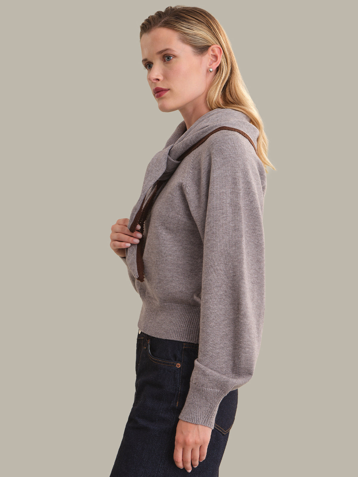 A woman in side profile wearing a light gray knit sweater with a brown shoulder strap and dark blue jeans, set against a solid light gray background.