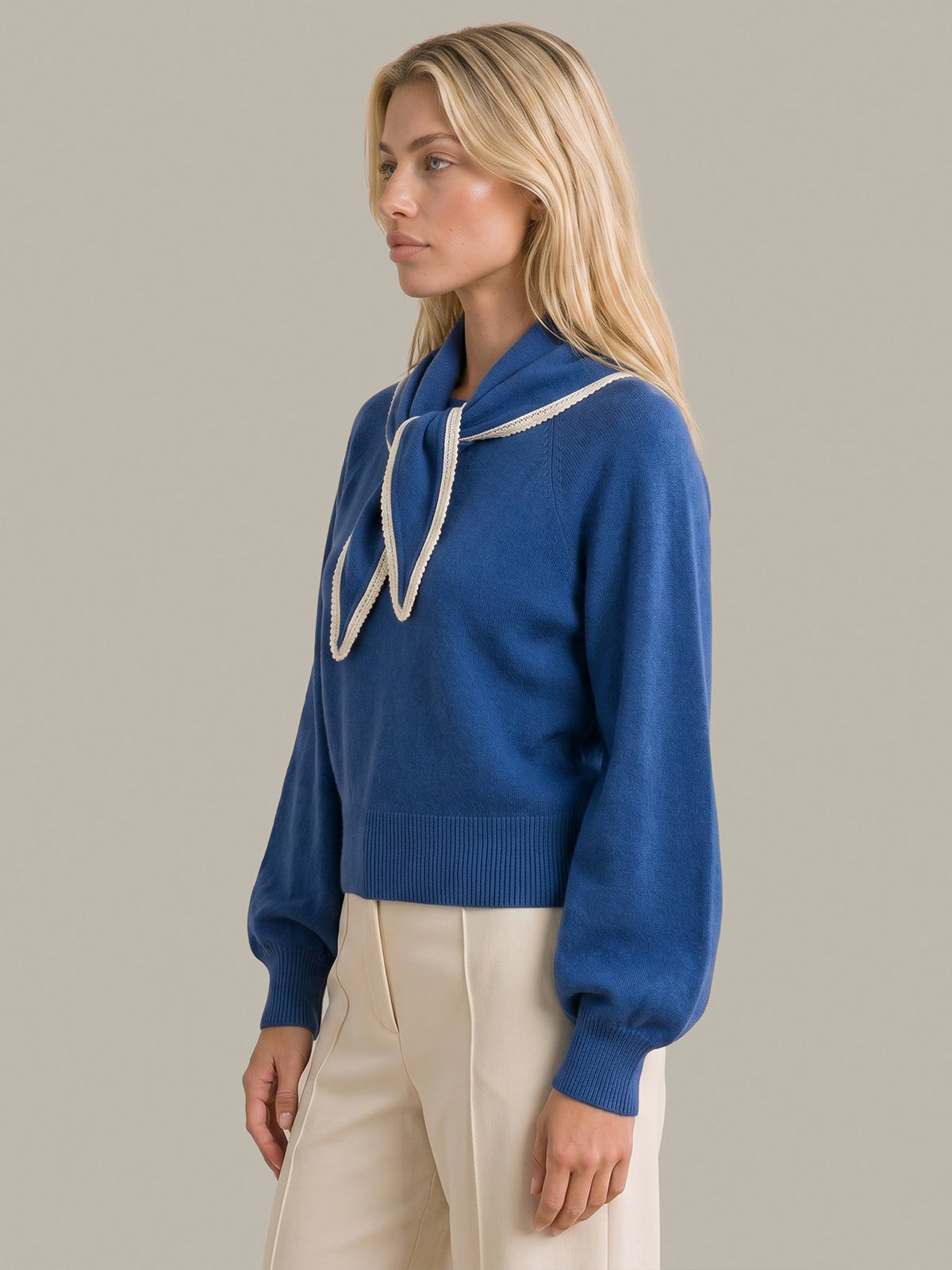 A blonde woman wearing a royal blue knit sweater with a high collar and cream rope details, paired with cream trousers, set against a solid light gray background.